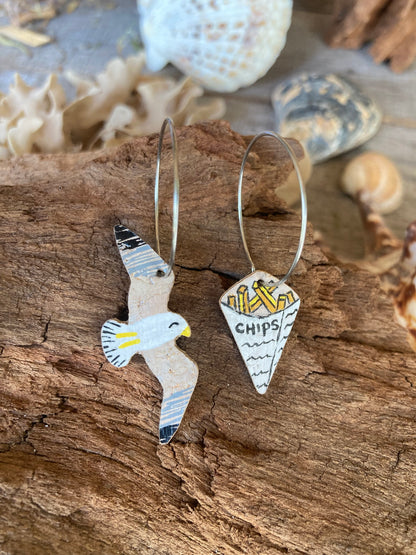 Seagull and chips mismatched cork earrings on silver hoops resting on driftwood with shells and coral — handpainted sustainable cork earrings handmade in UK