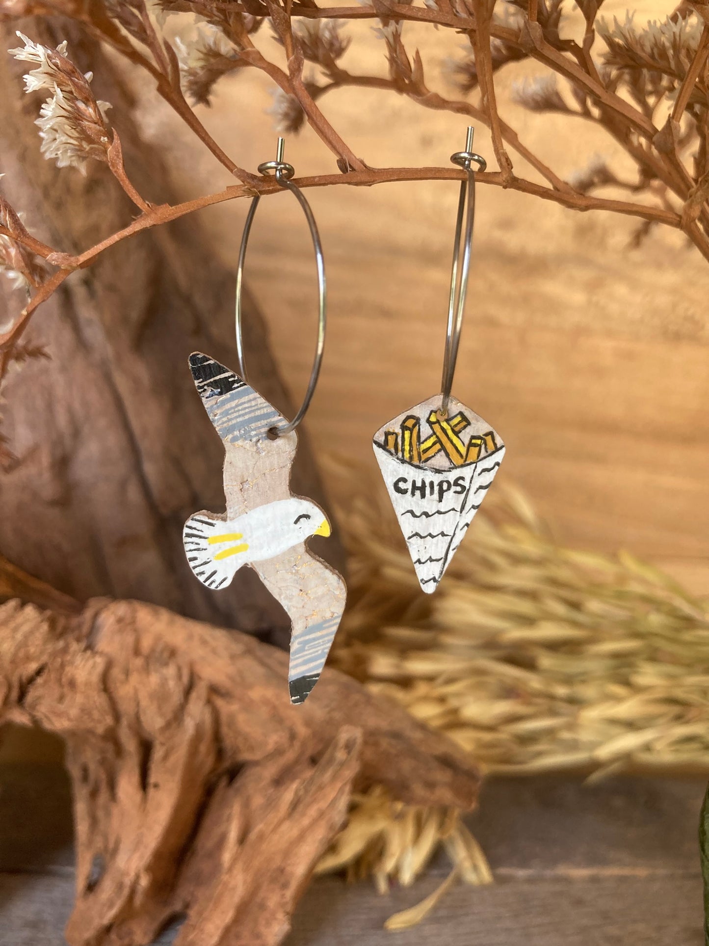 Seagull and chips cork earrings on silver hoops hanging from a dried russet botanical branch against driftwood — handpainted mismatched earrings handmade in Suffolk UK