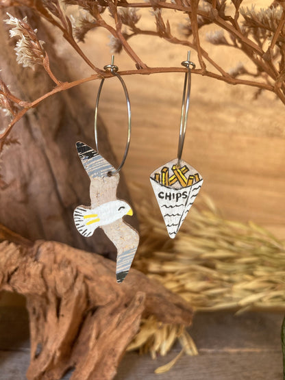 Seagull and chips cork earrings on silver hoops hanging from a dried russet botanical branch against driftwood — handpainted mismatched earrings handmade in Suffolk UK