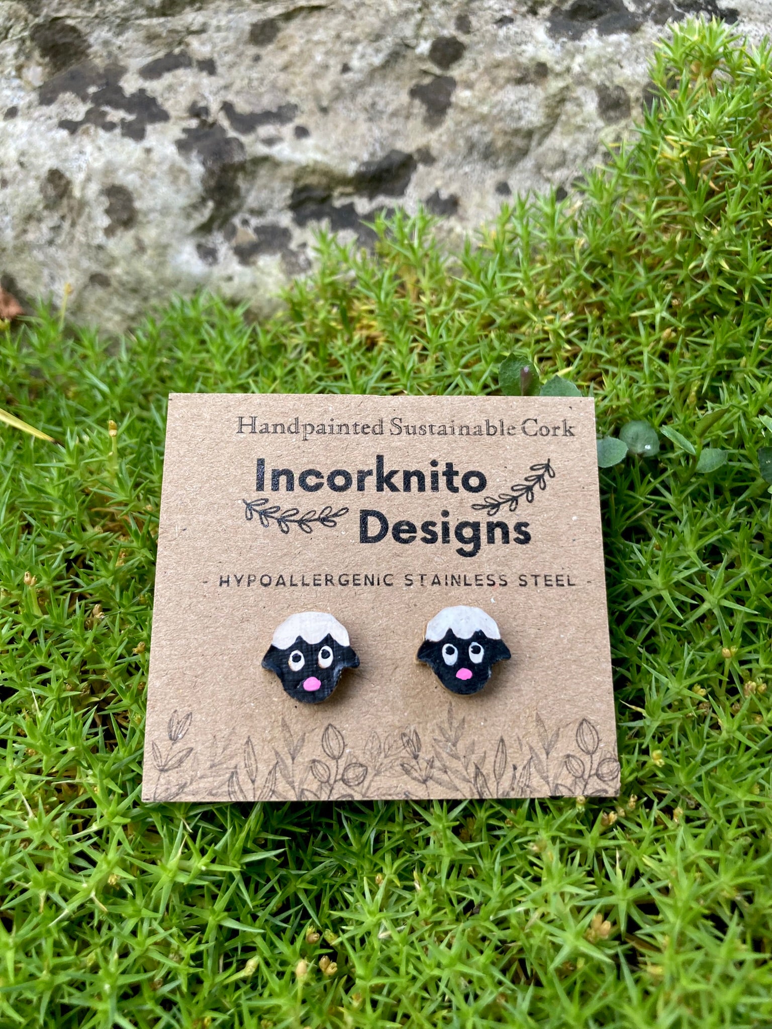 Handpainted sheep cork stud earrings on a branded card resting on green moss with a stone wall background