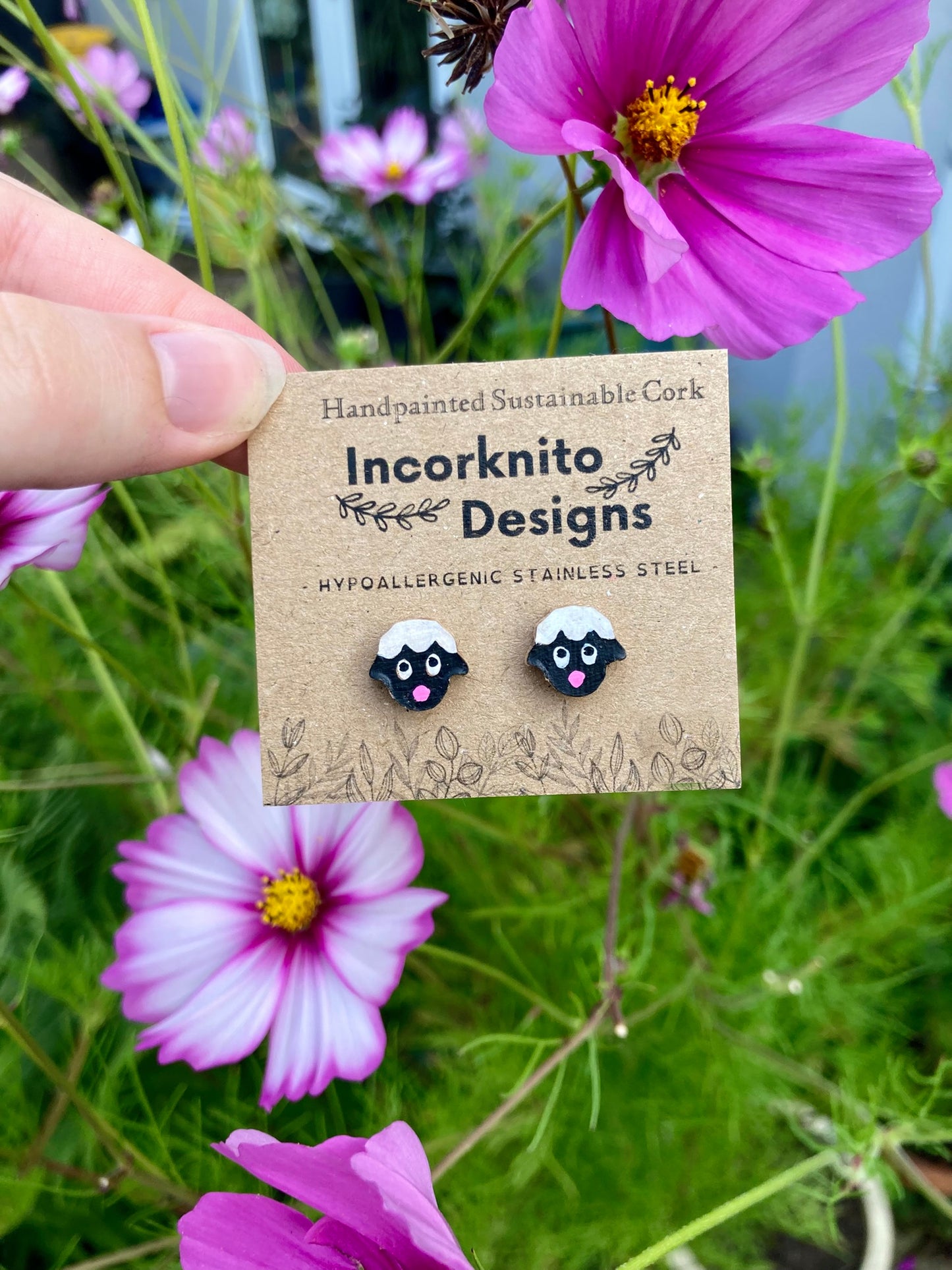 Sheep stud earrings on a branded card held in front of pink cosmos flowers in a garden setting
