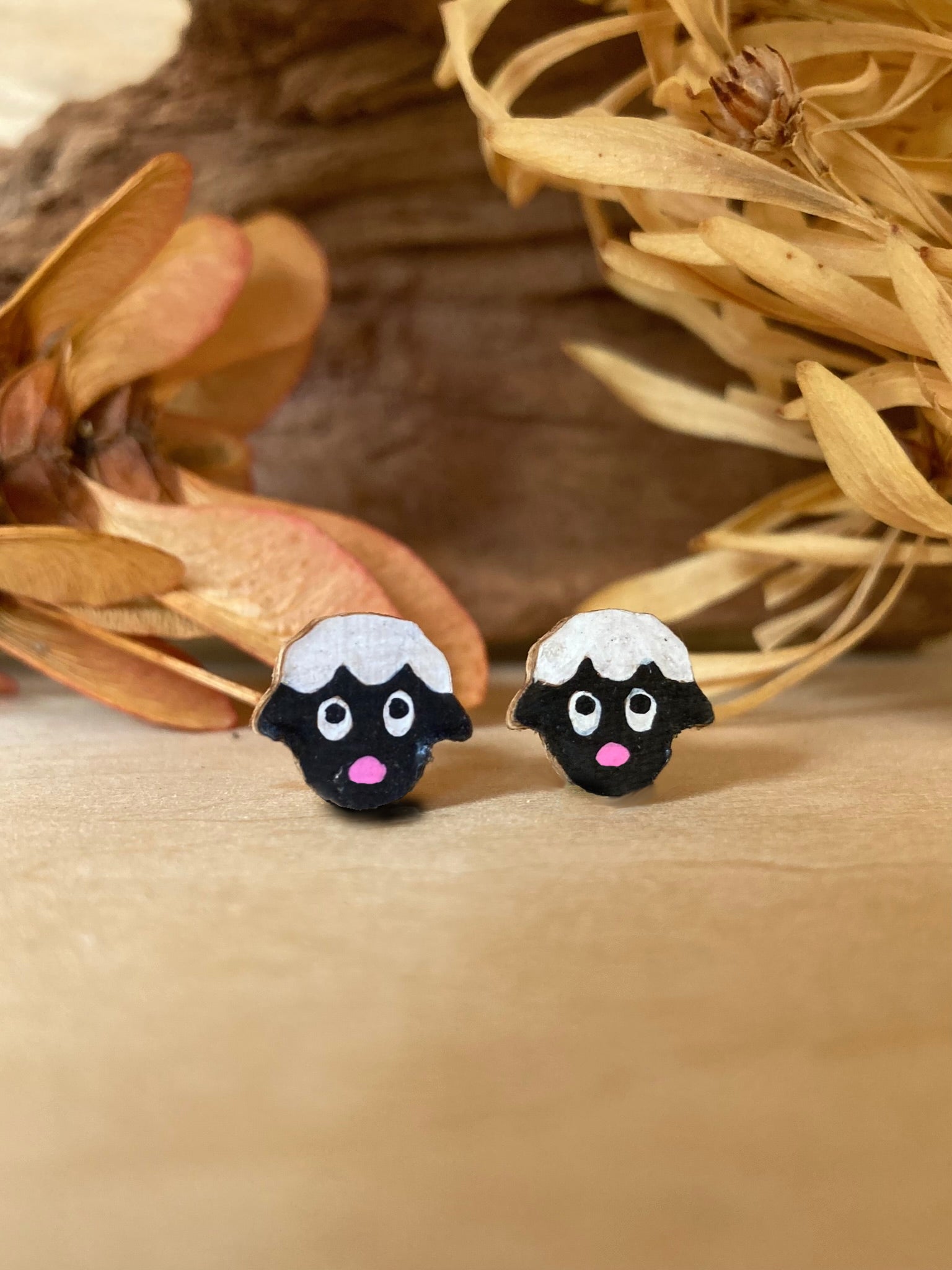Handpainted cork sheep stud earrings on a wooden surface with dried autumn leaves and driftwood