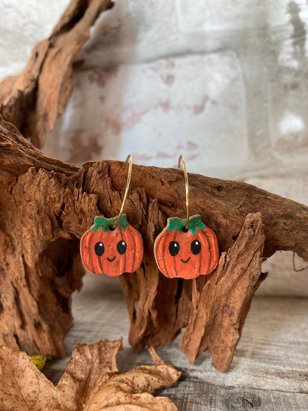 Orange pumpkin cork earrings hanging from textured bark on gold hoops, with green tops, black oval eyes and little curved smiles.