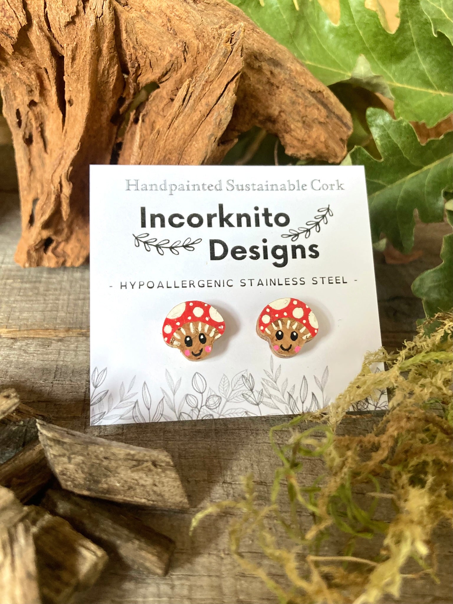 Smiley toadstool cork stud earrings on an Incorknito Designs branded card on a driftwood surface with moss and autumn leaves