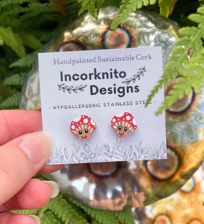 Smiley toadstool cork stud earrings on an Incorknito Designs branded card held against a fern and metal garden ornament
