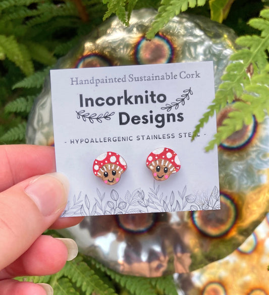 Smiley toadstool cork stud earrings on an Incorknito Designs branded card held against a fern and metal garden ornament
