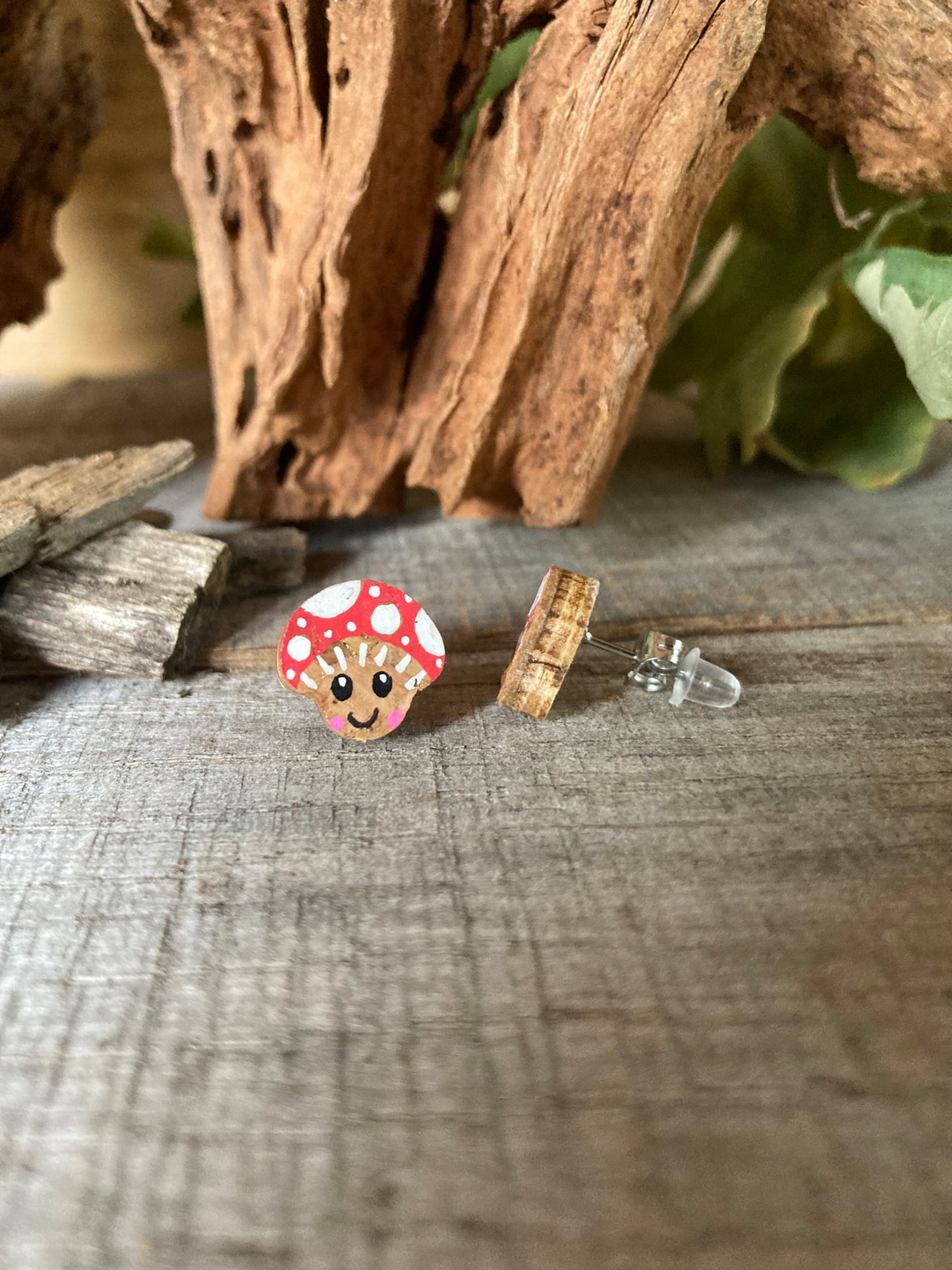 Smiley toadstool cork stud earrings on a driftwood surface with wild greenery. Side view of FSC wood with backing options