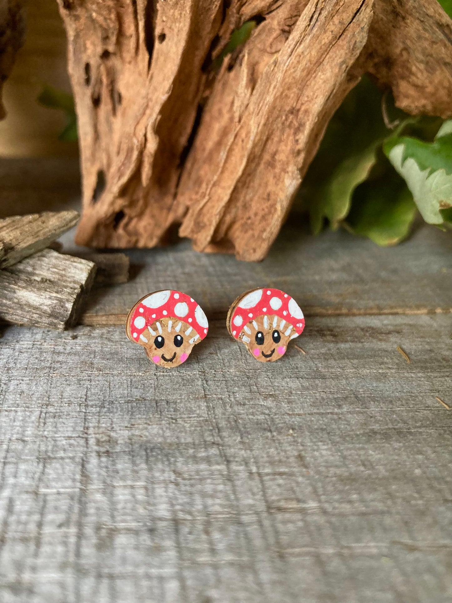 Handpainted smiley toadstool cork stud earrings with red cap, white polka dots and happy face on a driftwood surface
