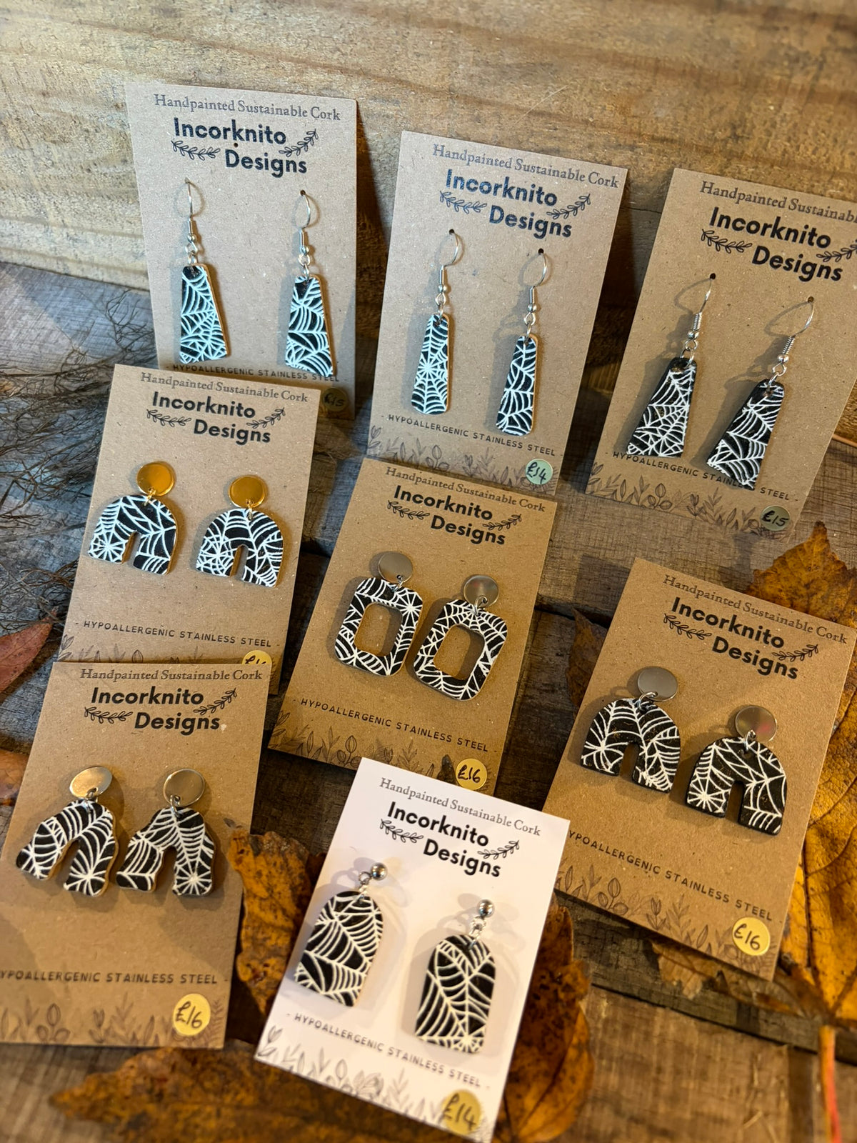 Collection of handpainted black cork earrings with white spider-web patterns, showing arch, rectangle and tapered designs with hooks and stud-top fastenings.