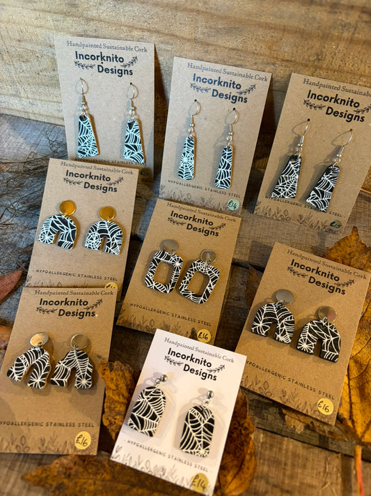 Collection of handpainted black cork earrings with white spider-web patterns, showing arch, rectangle and tapered designs with hooks and stud-top fastenings.