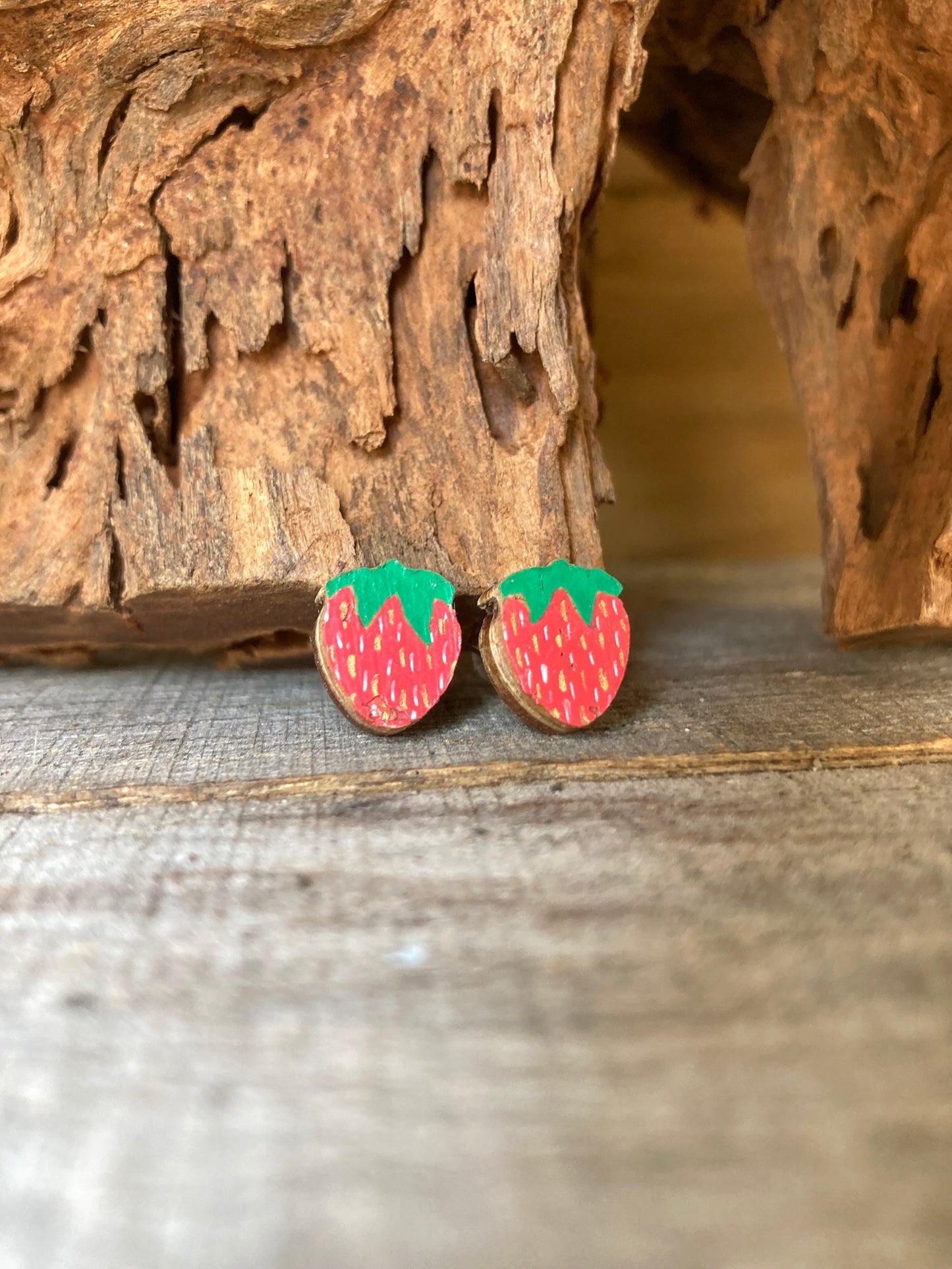 Handpainted strawberry cork stud earrings on a driftwood surface with rustic bark background. White and gold flecked detailing for the seeds and a vivid green cap