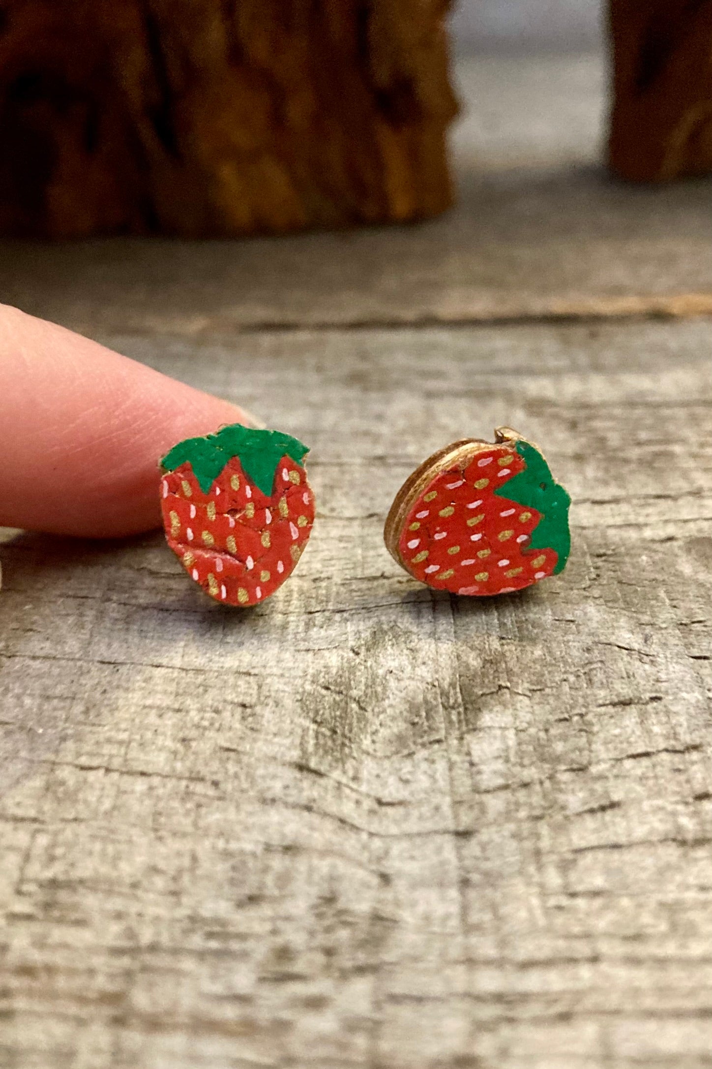 Handpainted strawberry cork stud earrings with red body, green leaf top and white seed detail on a driftwood surface