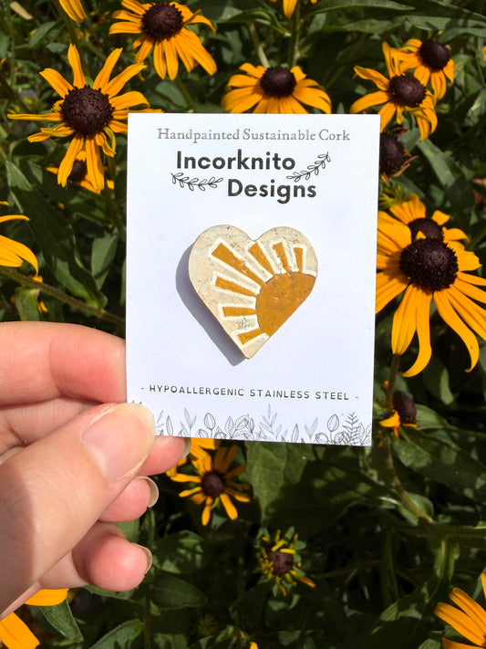 Sun heart cork pin badge on Incorknito Designs branded card held against yellow rudbeckia flowers — handpainted sunrise heart, sustainable cork badge handmade in Suffolk UK