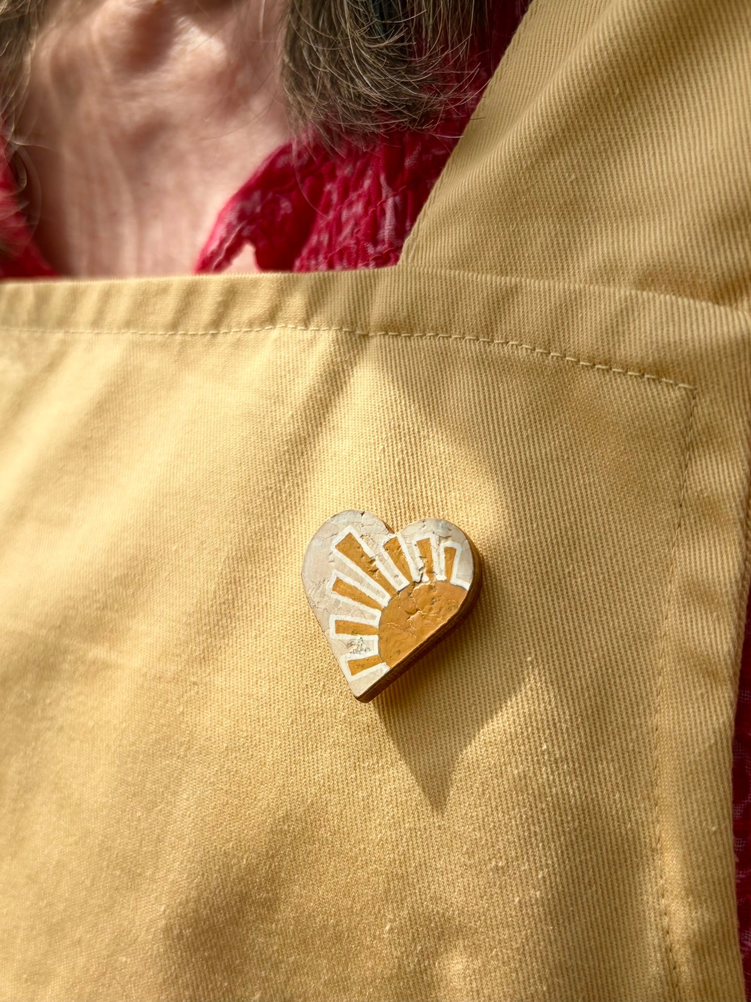 Sun heart cork pin badge pinned to a mustard yellow linen apron pocket — handpainted golden sunrise heart, sustainable cork badge