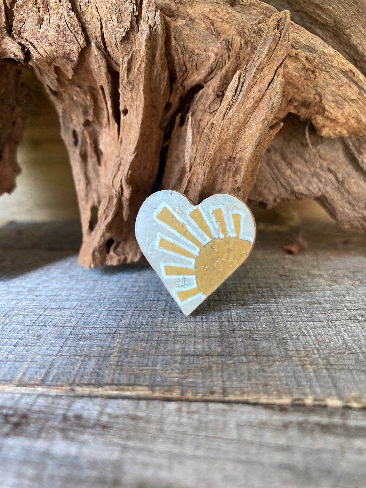 Handpainted sun cork pin badge propped against driftwood — heart-shaped badge with golden rising sun design, handmade in Suffolk UK