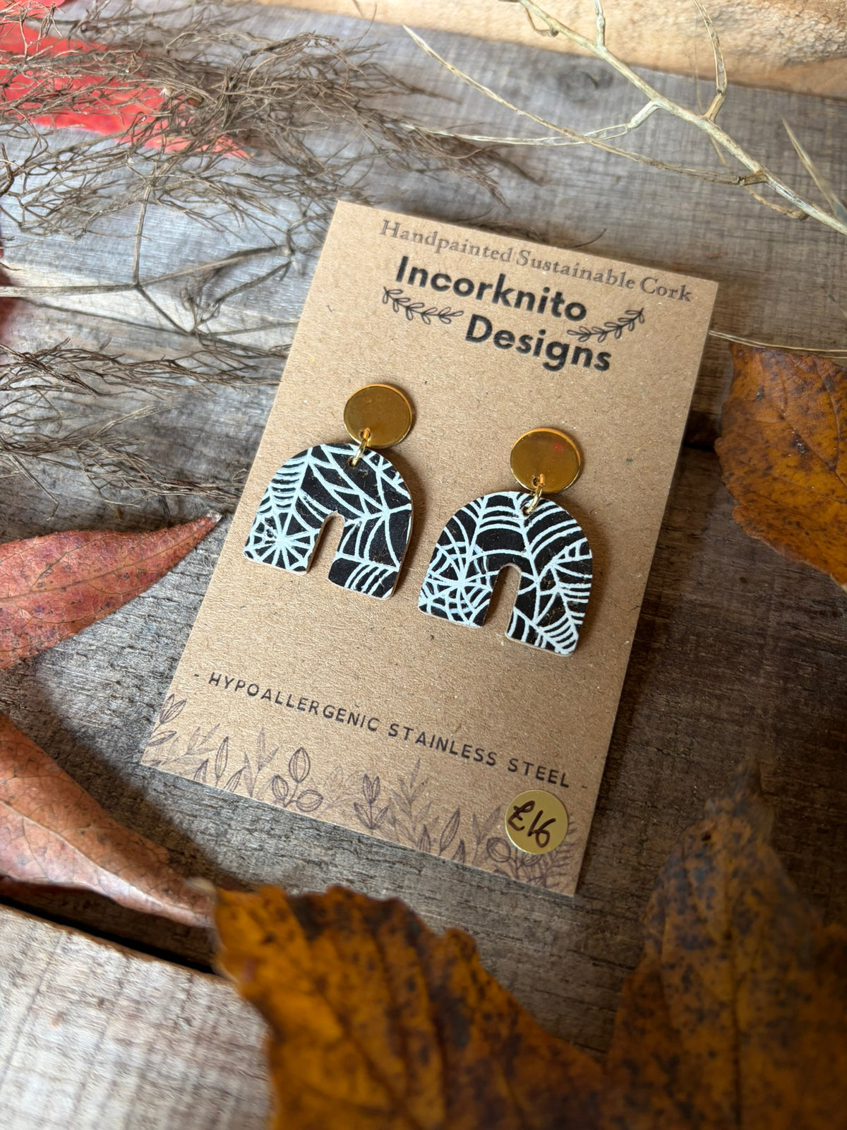 SW2 black cork arch earrings with gold disc stud tops and handpainted white cobwebs, styled on a display card among autumn leaves.