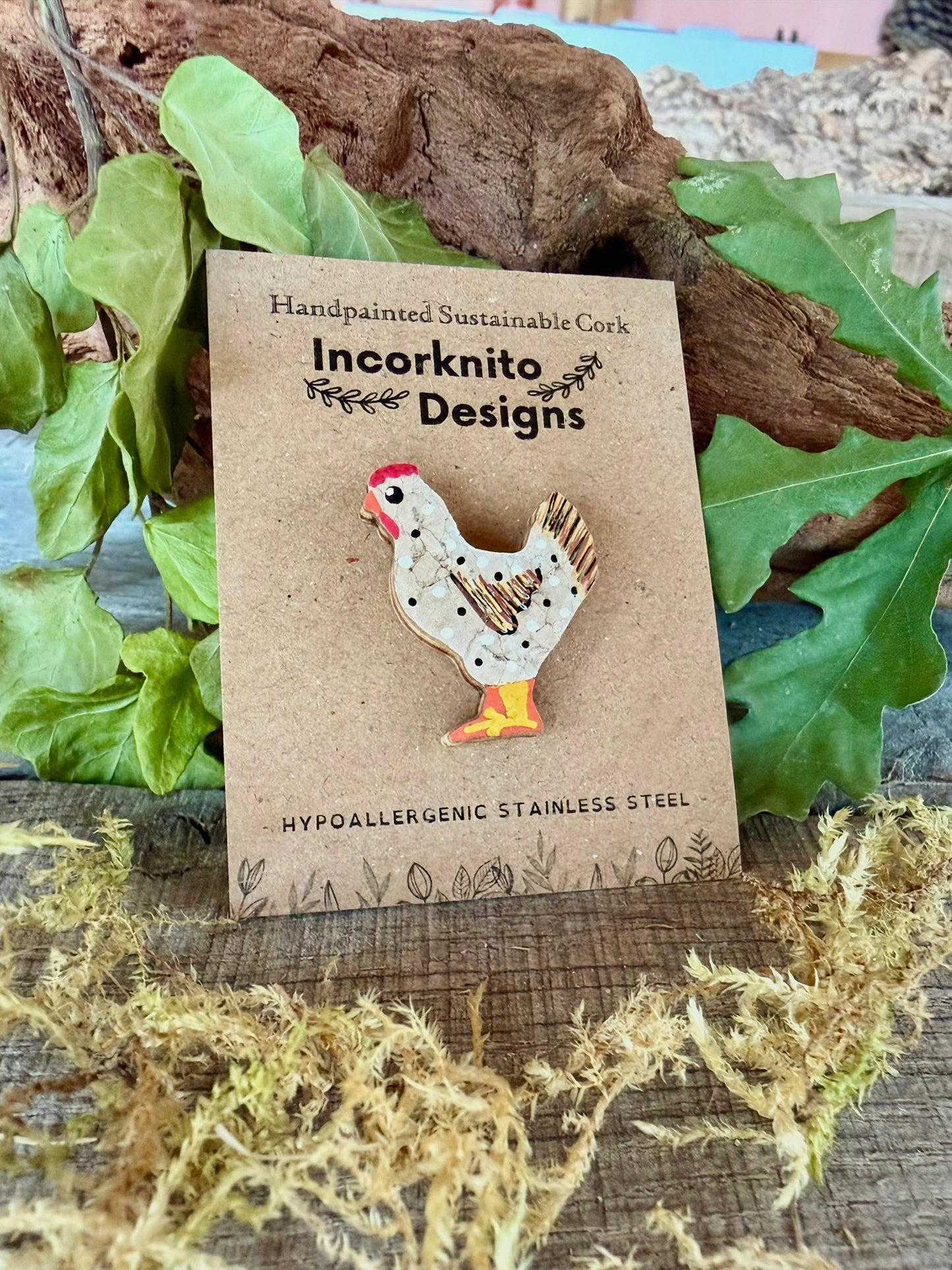 White chicken cork pin badge on Incorknito Designs branded card against driftwood, dried moss and oak leaves — handpainted sustainable cork badge