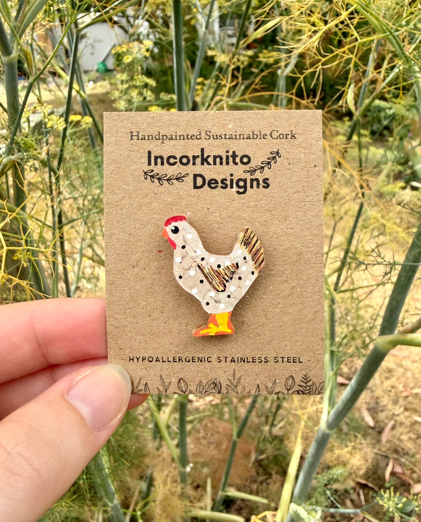 White chicken cork pin badge on Incorknito Designs branded card held against garden fennel — handpainted hen with polka dots, sustainable cork badge handmade in Suffolk UK