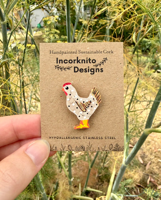 White chicken cork pin badge on Incorknito Designs branded card held against garden fennel — handpainted hen with polka dots, sustainable cork badge handmade in Suffolk UK