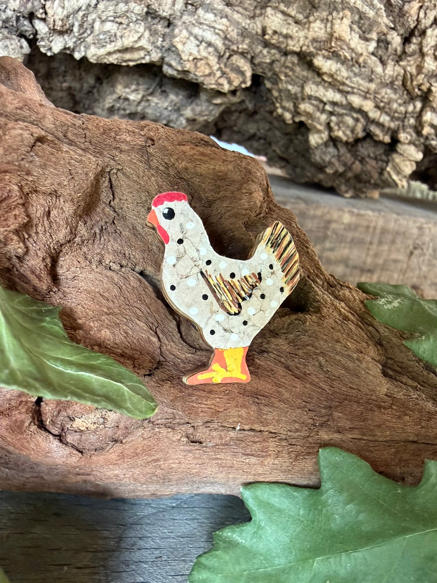 White chicken cork pin badge propped against driftwood with oak leaves — handpainted white hen with polka dots, red comb and golden tail feathers