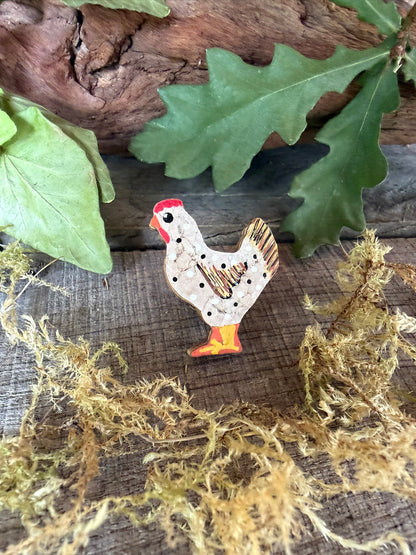 Handpainted white chicken cork pin badge face-on on weathered wood with dried moss and oak leaves — white hen with white and black polka dots, red comb and gold cork tail feathers