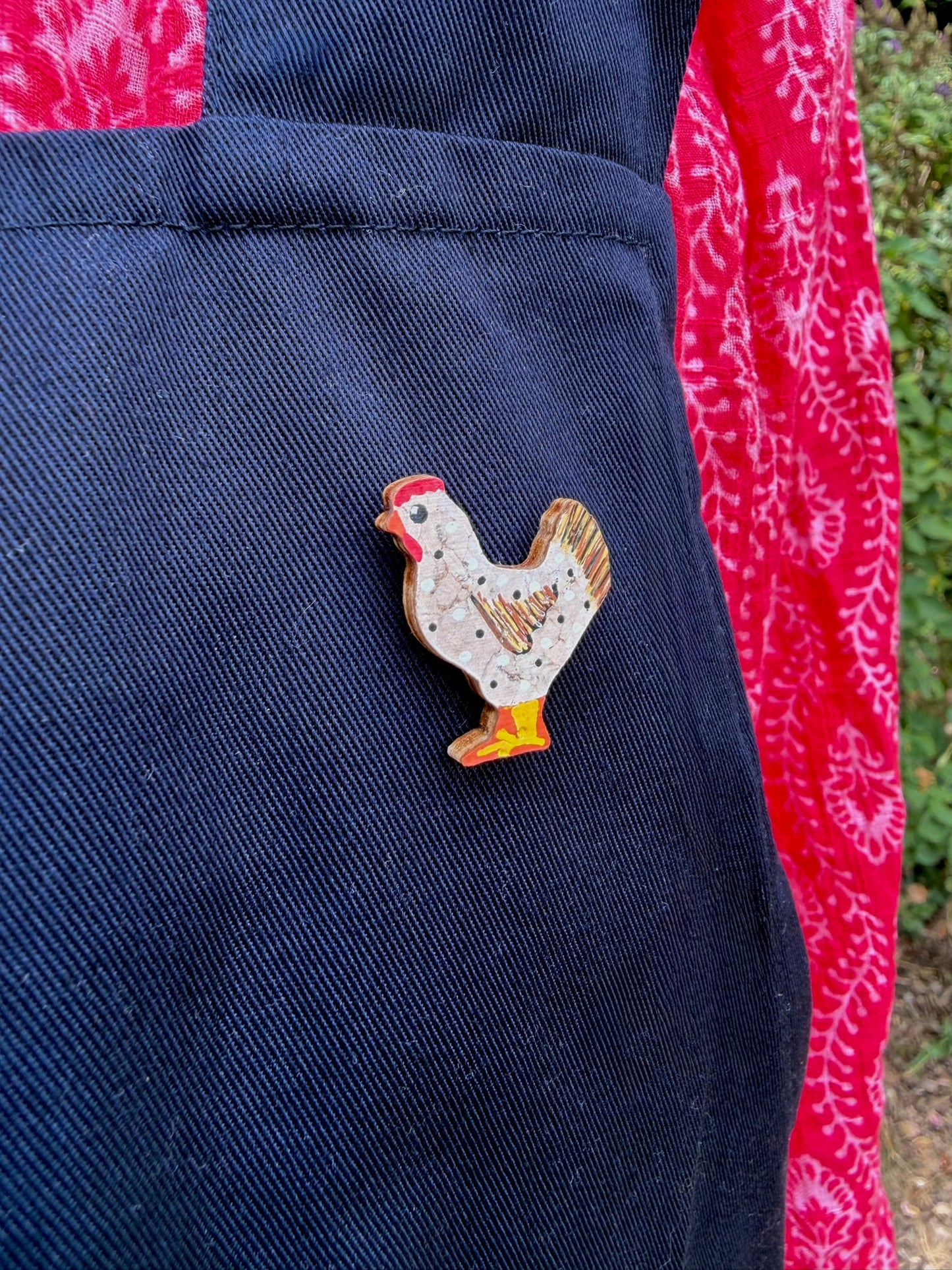 White chicken cork pin badge pinned to a navy linen apron pocket — handpainted hen with polka dots and golden tail feathers, sustainable cork badge
