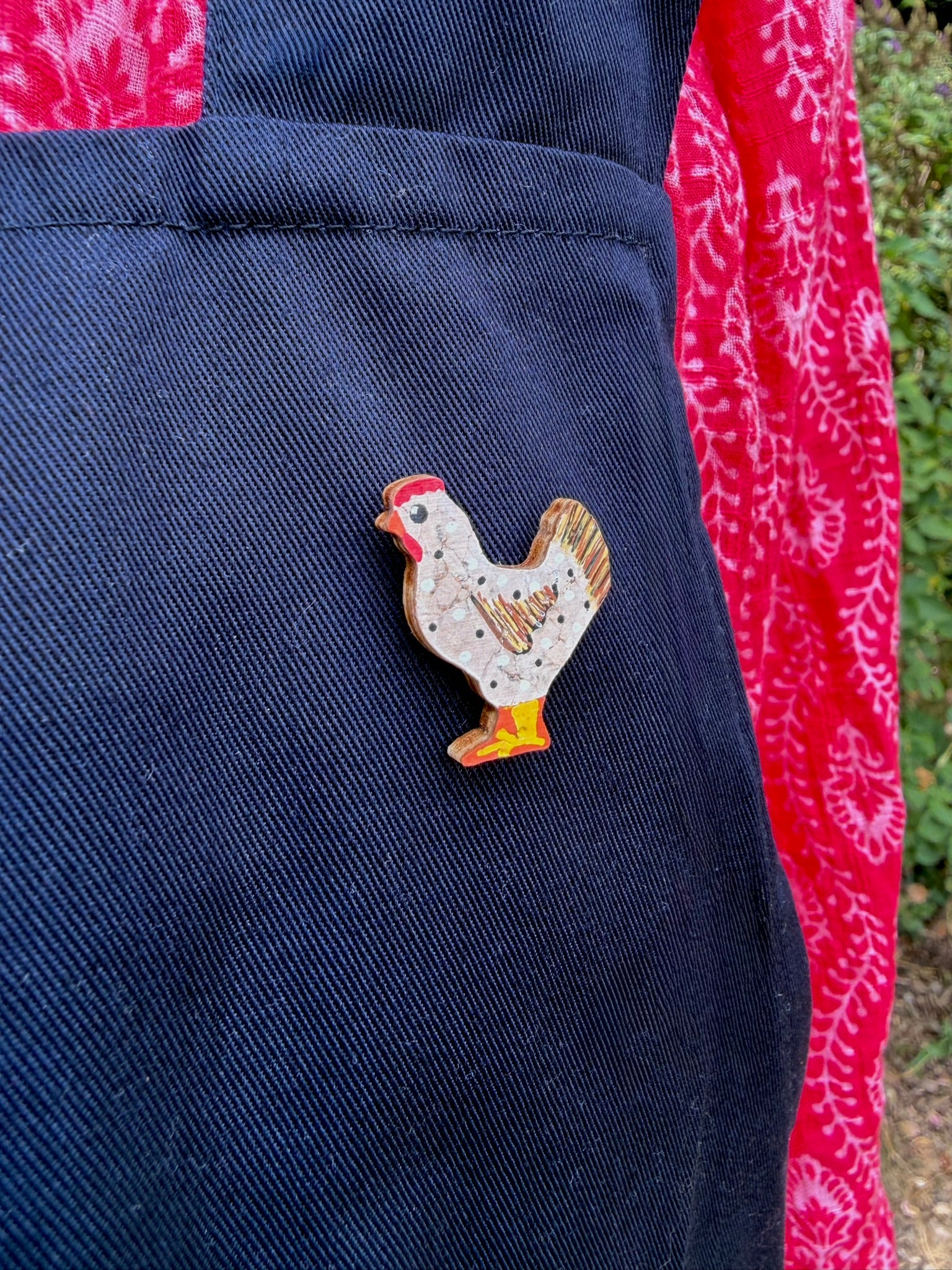White chicken cork pin badge pinned to a navy linen apron pocket — handpainted hen with polka dots and golden tail feathers, sustainable cork badge