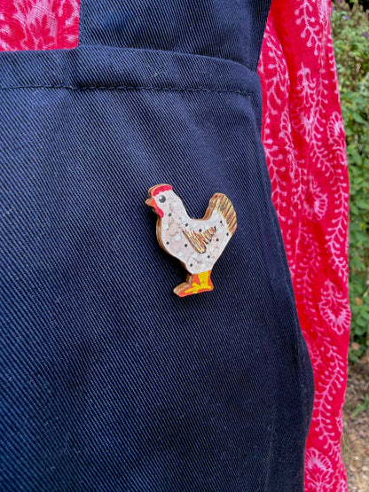 White chicken cork pin badge pinned to a navy linen apron pocket — handpainted hen with polka dots and golden tail feathers, sustainable cork badge